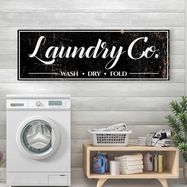 Puede incluir: Un letrero en blanco y negro con el texto "Laundry Co." y las palabras "Wash, Dry, Fold" debajo. El letrero est&aacute; colgado en una pared de madera blanca sobre una lavadora blanca.
