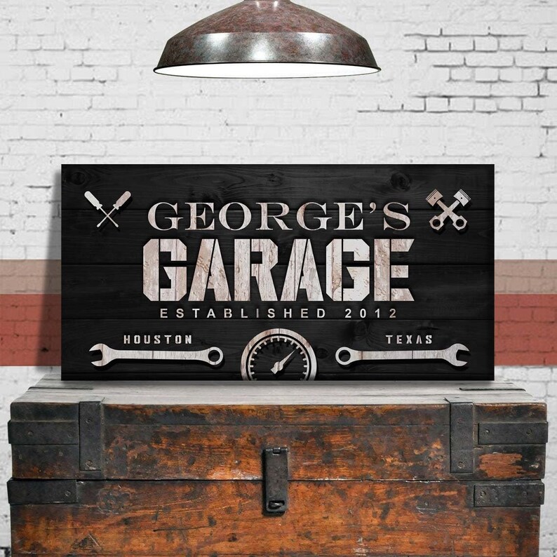 May include: A black wooden garage sign with the text "GEORGE'S GARAGE ESTABLISHED 2012". The sign includes images of tools and a speedometer. The sign is displayed on a wooden chest.