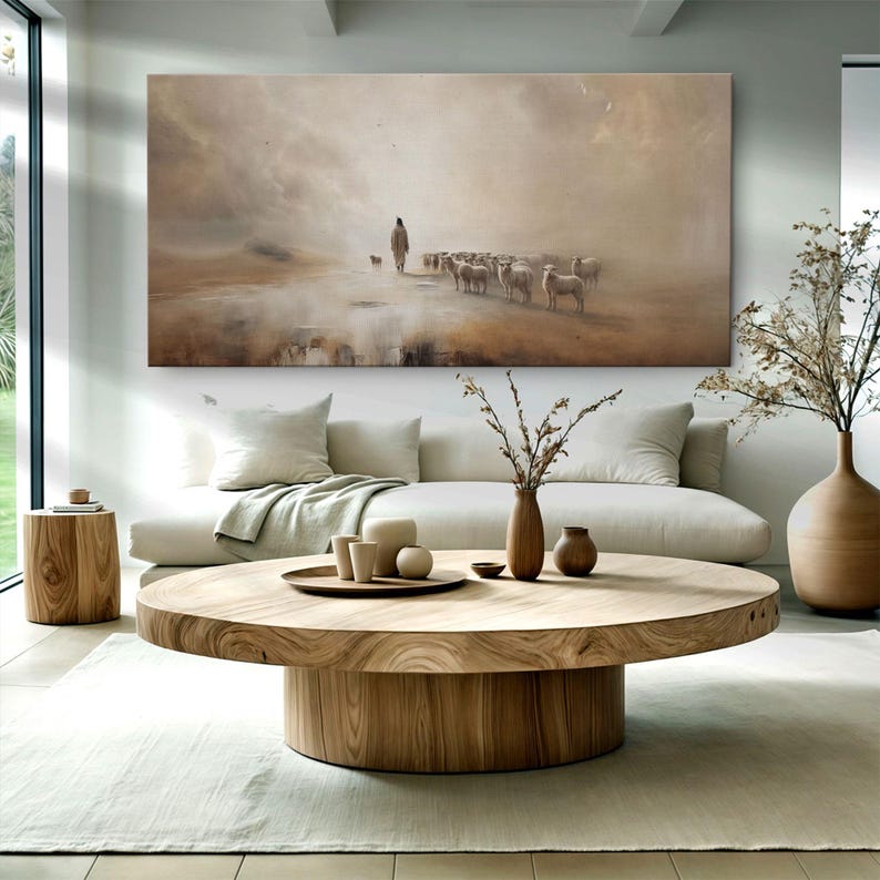 Puede incluir: Una gran obra de arte en lienzo rectangular que representa a un pastor y su reba&ntilde;o de ovejas en un paisaje en tonos sepia. La obra cuelga sobre un sof&aacute; blanco y una mesa de centro redonda de madera.