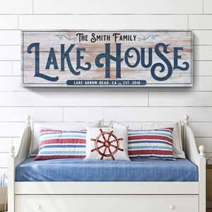 以下が含まれることがあります： 「LAKE HOUSE」の文字がネイビーブルーの筆記体で書かれた長方形の木製看板。その下に「LAKE ARROW HEAD, CA EST. 2010」と書かれています。看板の下には、ストライプのクッションと船の舵のクッションが付いたデイベッドがあります。