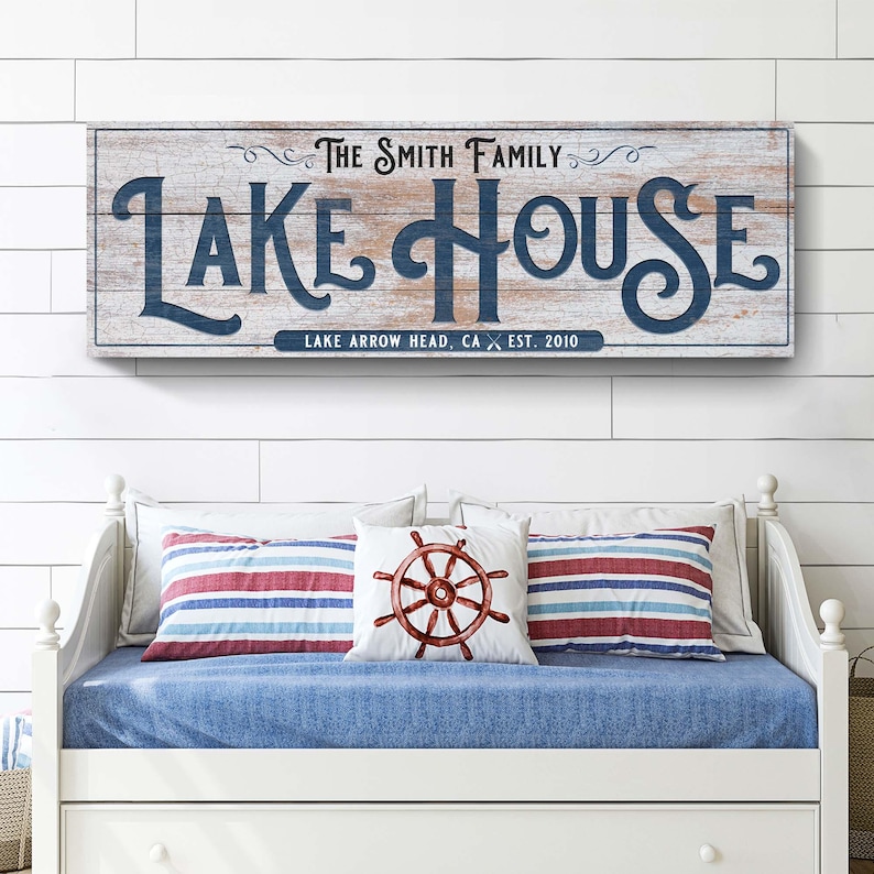 以下が含まれることがあります： 「LAKE HOUSE」の文字がネイビーブルーの筆記体で書かれた長方形の木製看板。その下に「LAKE ARROW HEAD, CA EST. 2010」と書かれています。看板の下には、ストライプのクッションと船の舵のクッションが付いたデイベッドがあります。