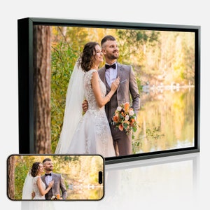 May include: A bride and groom pose for a wedding photo in a forest setting. The bride is wearing a white wedding dress and veil, and the groom is wearing a gray suit with a black bow tie. They are standing in front of a lake with trees and foliage in the background.