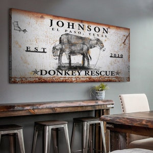 May include: Rustic metal sign with a distressed white background and brown rust accents. The sign features a black and white illustration of two donkeys, the text "JOHNSON EL PASO, TEXAS EST 2010 DONKEY RESCUE", and a star.