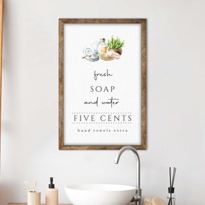 May include: A framed watercolor illustration of a bathroom sign with the text "fresh SOAP and water FIVE CENTS hand towels extra". The illustration includes a bar of soap, a bottle of liquid soap, a towel, and a potted plant.