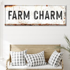 Puede incluir: Un letrero de metal r&uacute;stico con fondo blanco y letras negras que dice "FARM CHARM". El letrero tiene un aspecto desgastado y envejecido con manchas de &oacute;xido.