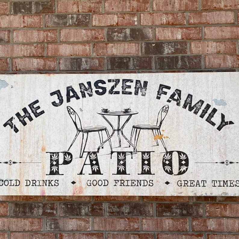 K&ouml;nnte beinhalten: Ein wei&szlig;es Schild mit schwarzem Text, auf dem "The Janszen Family Patio" steht, mit einer Zeichnung eines Tisches und zweier St&uuml;hle. Das Schild sagt auch "Cold Drinks, Good Friends, Great Times".
