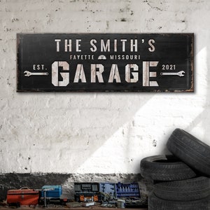 May include: Black wooden sign with white text that reads "The Smith's Garage Est. 2021 Fayette Missouri". The sign has a distressed look and features wrench icons on either side of the word "Garage".