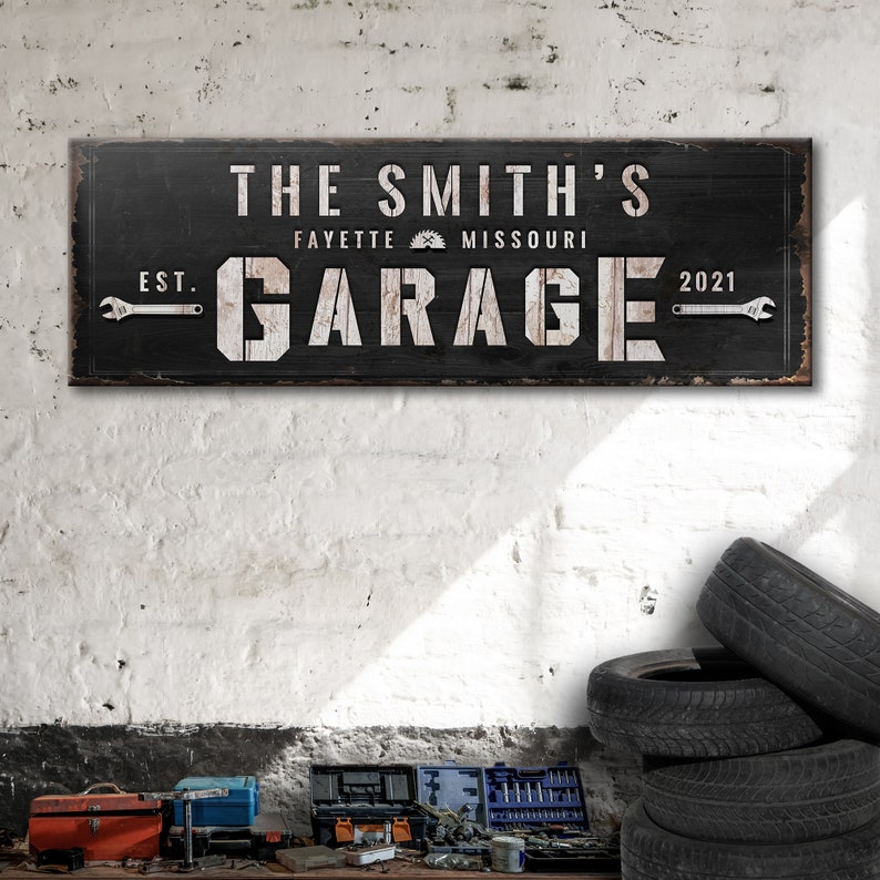May include: Black wooden sign with white text that reads "The Smith's Garage Est. 2021 Fayette Missouri". The sign has a distressed look and features wrench icons on either side of the word "Garage".