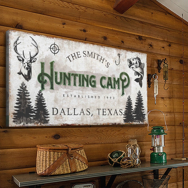 Puede incluir: Un cartel r&uacute;stico blanco con texto verde que dice "The Smith's Hunting Camp Established 1993 Dallas, Texas". El cartel presenta una cabeza de ciervo, un anzuelo de pesca y dos pinos.
