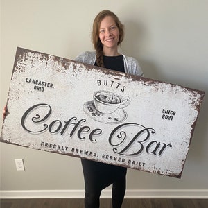 May include: A white sign with a rustic, distressed look, featuring the text "Butts Coffee Bar" in a stylized font. The sign also includes the text "Lancaster, Ohio" and "Since 2021". A black and white illustration of a coffee cup with steam rising from it is featured on the sign.