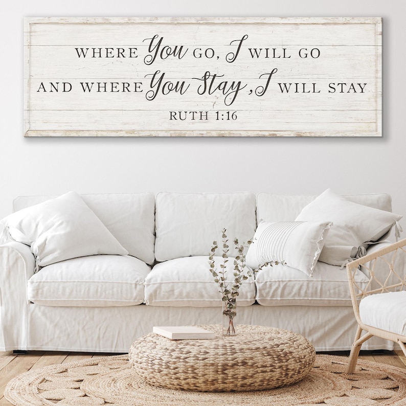 K&ouml;nnte beinhalten: Ein rechteckiges Holzschild mit dem Text "WHERE You GO, I WILL GO AND WHERE You Stay, I WILL STAY RUTH 1:16" in schwarzer Schrift. Das Schild ist &uuml;ber einem wei&szlig;en Sofa mit mehreren Kissen angebracht. Ein runder geflochtener Couchtisch und ein Teppich befinden sich vor dem Sofa.