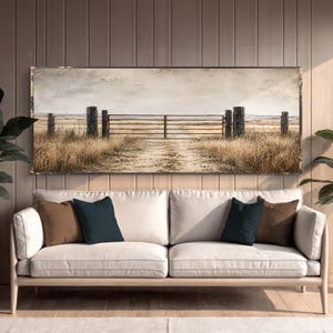 May include: A rectangular landscape wall art print featuring a rustic metal gate set between weathered wooden fence posts in a dry, golden-brown field under a pale, overcast sky, displayed above a light-colored sofa with brown and dark blue pillows.