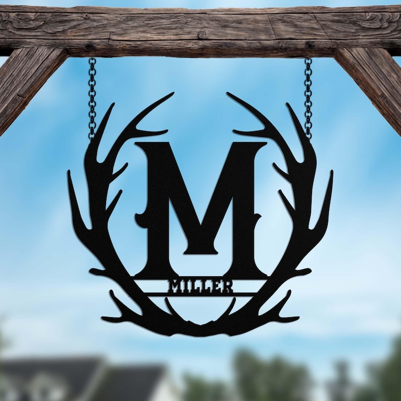May include: Black metal sign featuring a large letter M at its centre, encircled by a wreath of antlers. The name "Miller" is inscribed beneath the letter.