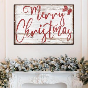 Puede incluir: Un letrero de madera blanco con letras rojas que dice "Merry Christmas" con un lazo rojo en la esquina superior derecha. El letrero tiene un aspecto desgastado y est&aacute; cubierto de copos de nieve blancos.