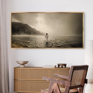 Pu&ograve; includere: Stampa artistica incorniciata in tonalit&agrave; seppia raffigurante una figura che cammina nell'acqua. Montagne e un cielo nuvoloso fanno da sfondo. L'opera d'arte &egrave; esposta sopra un mobile in legno con una trama intrecciata.