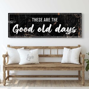 May include: Black and white sign with distressed metal texture. The sign reads "These are the good old days". The sign is hanging above a wooden bench with white pillows.