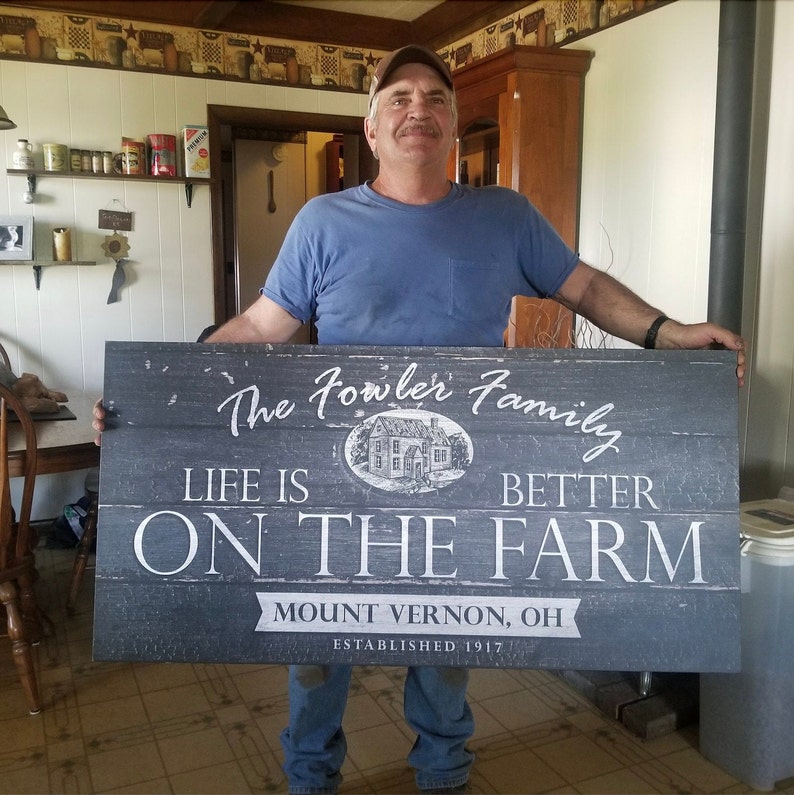 May include: A black and white sign with the text "The Fowler Family Life is Better On The Farm Mount Vernon, OH Established 1917". A man is holding the sign in front of him.