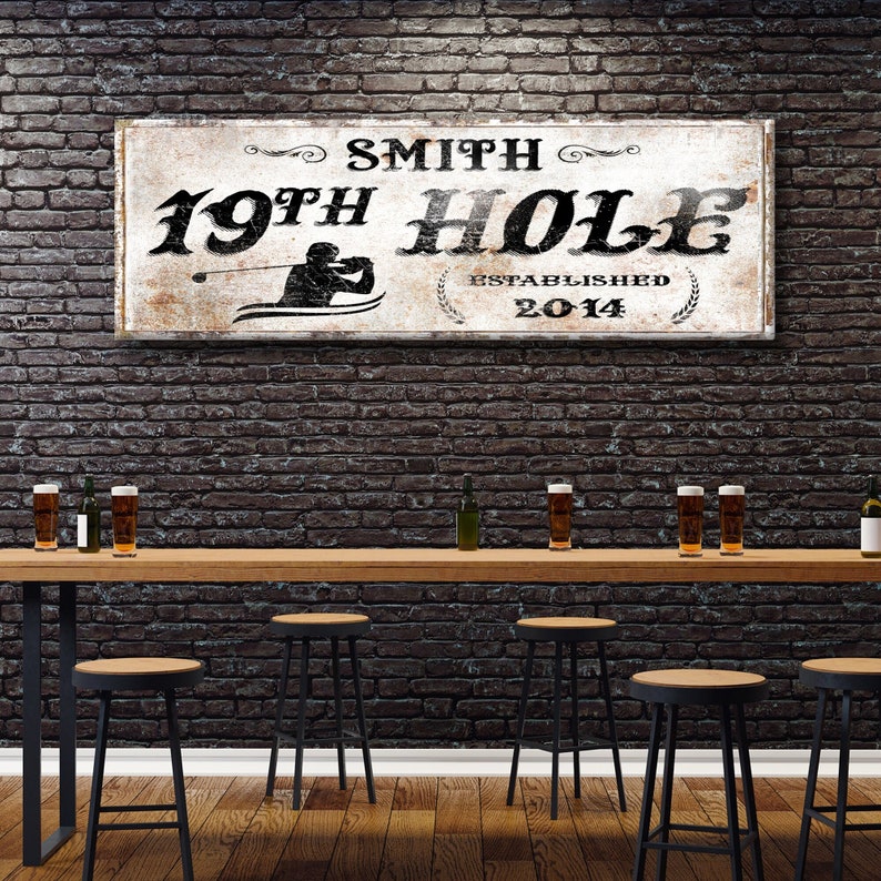 Puede incluir: Un letrero de madera r&uacute;stico con pintura blanca y letras negras. El letrero dice "Smith 19th Hole Established 2014" y presenta una silueta de un golfista balanceando un palo. El letrero est&aacute; colgado en una pared de ladrillo sobre una barra con taburetes.