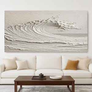 May include: A large, textured, off-white wave art piece. The artwork depicts a breaking wave with detailed ridges and foam, creating a three-dimensional effect. The piece is mounted above a white sofa and dark wood coffee table.