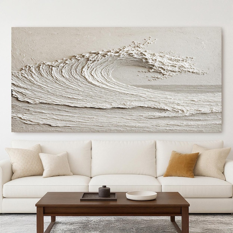 May include: A large, textured, off-white wave art piece. The artwork depicts a breaking wave with detailed ridges and foam, creating a three-dimensional effect. The piece is mounted above a white sofa and dark wood coffee table.