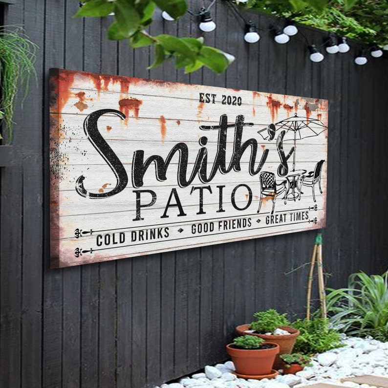 K&ouml;nnte beinhalten: Rustikales wei&szlig;es Holzschild mit schwarzem Text, der "Smith's Patio Est. 2020 Cold Drinks Good Friends Great Times" lautet. Das Schild hat einen abgenutzten Look mit rostigen braunen Akzenten und eine Schwarzwei&szlig;illustration eines Patio-Sets mit einem Sonnenschirm.