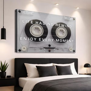 May include: A clear acrylic wall art piece shaped like a cassette tape. The artwork features black tape reels and the text "LIFE HAS NO REWIND ENJOY EVERY MOMENT" in white. The piece is mounted above a bed.
