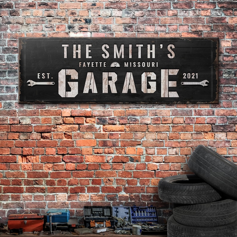 May include: Black wooden sign with white text that reads "The Smith's Garage Est. 2021 Fayette Missouri". The sign has a saw blade icon and wrench graphics.