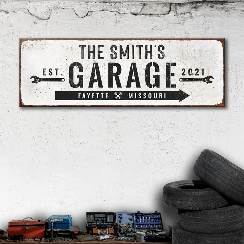 Puede incluir: Un letrero de metal blanco con texto negro que dice "The Smith's Garage Est. 2021 Fayette Missouri" con iconos de llave inglesa a cada lado del texto.