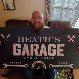 May include: A black wooden sign with the text "HEATH'S GARAGE BAR & GRILL" in white lettering. The sign also includes the text "DENTON TEXAS" and various mechanic-themed graphics, such as wrenches and crossed tools.