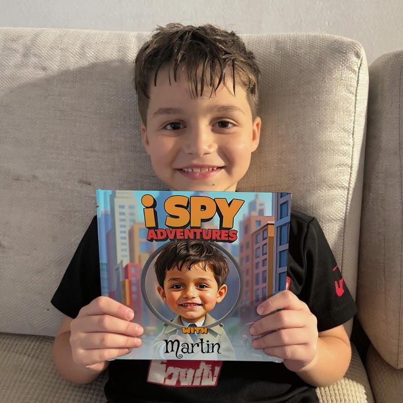 May include: A young boy smiles while holding a book titled "i SPY ADVENTURES" with a cartoon illustration of himself. The book cover features a cityscape background and the name "Martin" below the illustration.