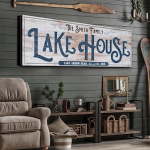 以下が含まれることがあります： ネイビーブルーの筆記体で「LAKE HOUSE」と書かれた木製の看板。その下に「LAKE ARROW HEAD, CA EST. 2010」と書かれています。看板は灰色の壁に掛けられており、オールやその他の航海をテーマにした装飾が施されています。