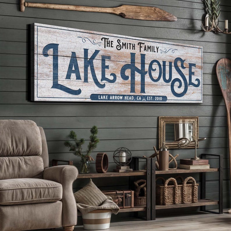 以下が含まれることがあります： ネイビーブルーの筆記体で「LAKE HOUSE」と書かれた木製の看板。その下に「LAKE ARROW HEAD, CA EST. 2010」と書かれています。看板は灰色の壁に掛けられており、オールやその他の航海をテーマにした装飾が施されています。