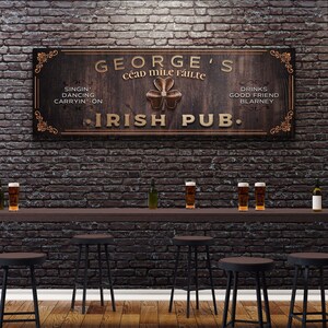 Puede incluir: Un letrero de madera con el texto "GEORGE'S C&eacute;ad Mile F&aacute;ilte IRISH PUB."  El letrero tiene un dise&ntilde;o de tr&eacute;bol y el texto "SINGIN' DANCING CARRYIN-ON" y "DRINKS GOOD FRIEND BLARNEY" a cada lado del tr&eacute;bol.