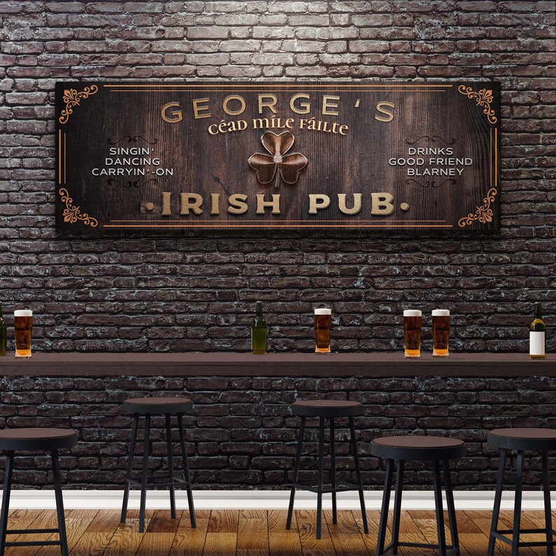 Puede incluir: Un letrero de madera con el texto "GEORGE'S C&eacute;ad Mile F&aacute;ilte IRISH PUB."  El letrero tiene un dise&ntilde;o de tr&eacute;bol y el texto "SINGIN' DANCING CARRYIN-ON" y "DRINKS GOOD FRIEND BLARNEY" a cada lado del tr&eacute;bol.