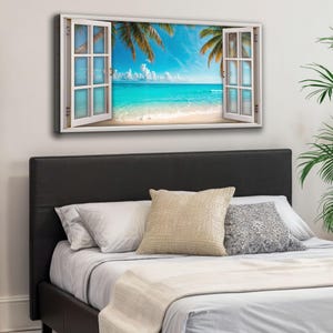 May include: A framed canvas print depicting a tropical beach scene viewed through a white window. The artwork shows a turquoise ocean, white sand, palm trees, and a bright blue sky with fluffy clouds. The print is mounted above a bed with pillows.