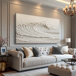 May include: A large, textured, neutral-toned artwork depicting a breaking ocean wave. The wave is the focal point, with detailed ridges and a foamy crest. The artwork hangs above a cream-colored tufted sofa with accent pillows, creating a serene living room setting.