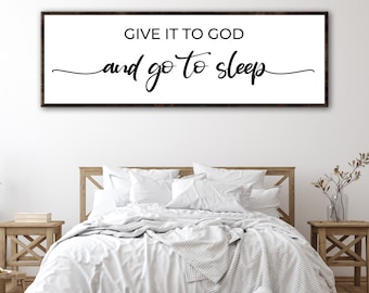 May include: A white rectangular sign with black text that reads "Give it to God and go to sleep". The sign is hanging above a white bed with white bedding and pillows. The bed is in a bedroom with wooden nightstands and a vase with dried branches.