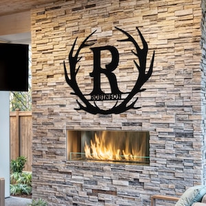 May include: Black metal wall decor featuring a large letter R surrounded by a wreath of antlers. The name "Robinson" is written below the letter. The decor is mounted on a stone wall above a fireplace.