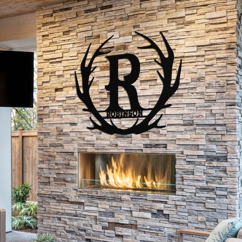 May include: Black metal wall decor featuring a large letter R surrounded by a wreath of antlers. The name "Robinson" is written below the letter. The decor is mounted on a stone wall above a fireplace.