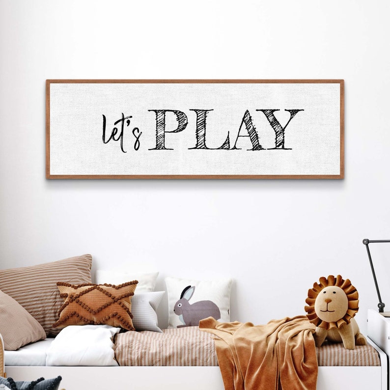 May include: A white canvas sign with a brown wood frame, featuring the text "Let's Play" in black script font.