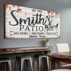 K&ouml;nnte beinhalten: Ein wei&szlig;es Holzschild mit schwarzem Text, der "Smith's Patio Est. 2020 Cold Drinks Good Friends Great Times Dallas, Texas" lautet. Das Schild hat einen abgenutzten Look mit rostigen Akzenten. Es gibt eine Schwarzwei&szlig;illustration eines Patio-Sets mit einem Sonnenschirm.