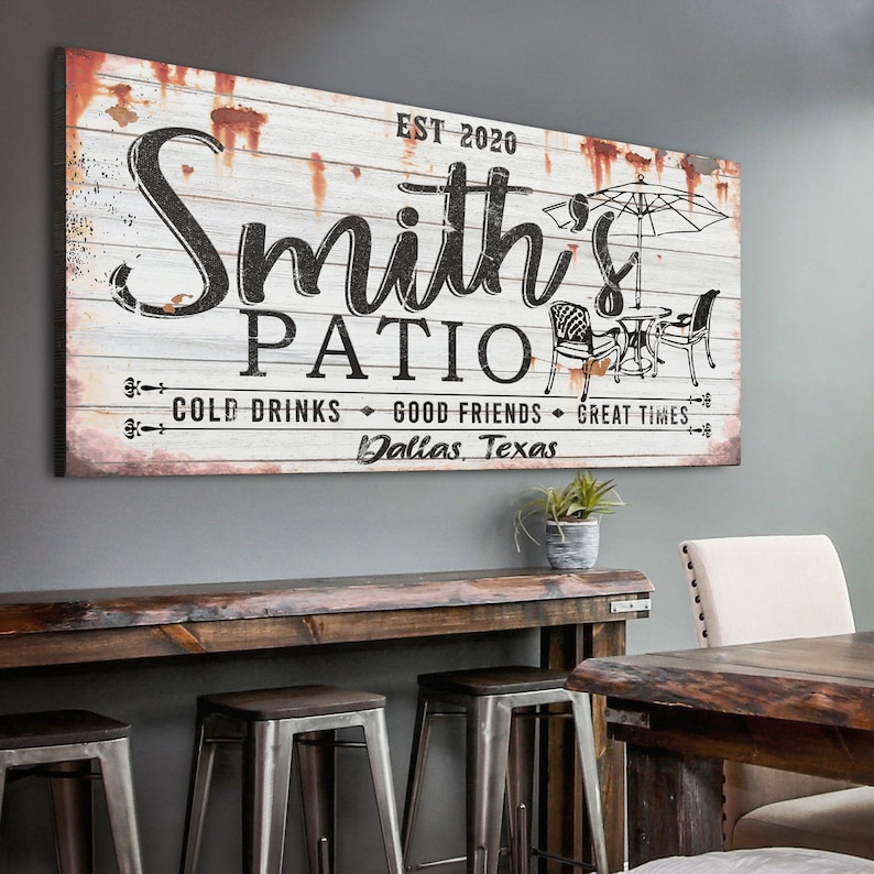 K&ouml;nnte beinhalten: Ein wei&szlig;es Holzschild mit schwarzem Text, der "Smith's Patio Est. 2020 Cold Drinks Good Friends Great Times Dallas, Texas" lautet. Das Schild hat einen abgenutzten Look mit rostigen Akzenten. Es gibt eine Schwarzwei&szlig;illustration eines Patio-Sets mit einem Sonnenschirm.