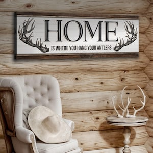 May include: A rustic wooden sign with the text "HOME IS WHERE YOU HANG YOUR ANTLERS" and two stylized antlers on either side of the text.