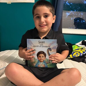 May include: A young person is sitting on a bed, holding a book titled "Sean in the World of Dinosaurs." The book cover features an illustration of a child and a dinosaur. The person is wearing a black shirt and shorts.