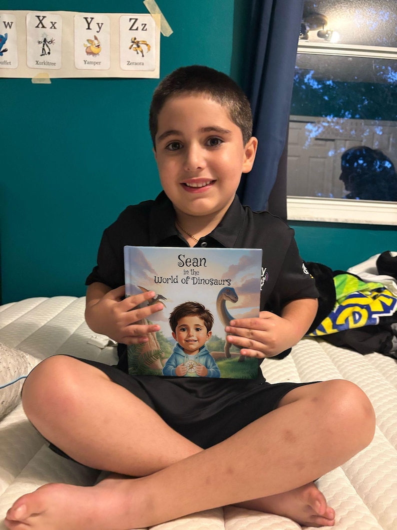 May include: A young person is sitting on a bed, holding a book titled "Sean in the World of Dinosaurs." The book cover features an illustration of a child and a dinosaur. The person is wearing a black shirt and shorts.