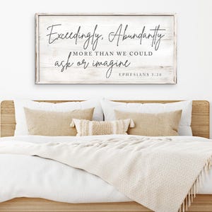 May include: White wooden sign with the text "Exceedingly Abundantly More Than We Could Ask Or Imagine" and the scripture reference "Ephesians 3:20". The sign is hanging above a bed with white bedding and tan pillows.