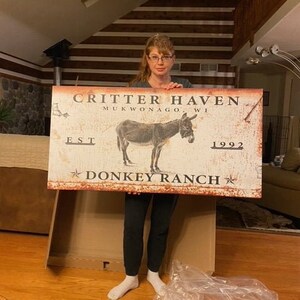 May include: A white sign with rusty edges featuring a donkey illustration and the text "Critter Haven Mukwonago, WI Est 1992 Donkey Ranch".