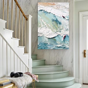 May include: An abstract oil painting of ocean waves with two surfers. The artwork features textured brushstrokes in shades of blue, white, and beige, creating a dynamic and energetic coastal scene. The painting is displayed on a marble wall.