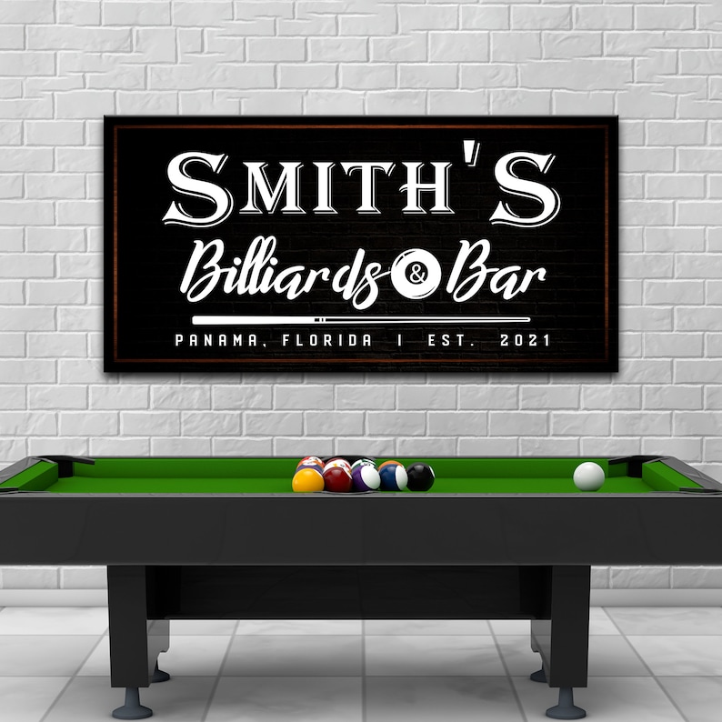 May include: A black and white sign with the text "Smith's Billiards & Bar" in a vintage style font. The sign also includes the text "Panama, Florida | Est. 2021". The sign is hanging on a white brick wall above a green pool table.