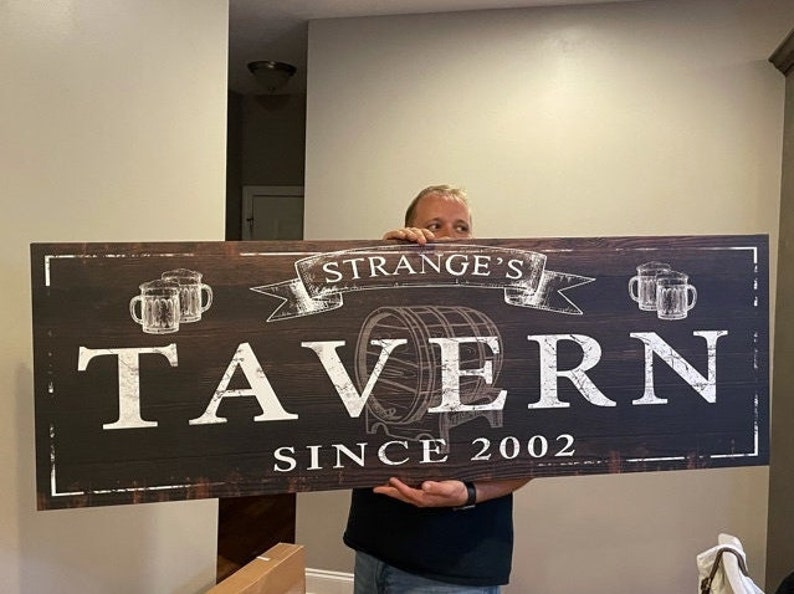 K&ouml;nnte beinhalten: Ein braunes Holzschild mit wei&szlig;em Text, der "Strange's Tavern Since 2002" lautet. Das Schild zeigt ein Fass, zwei Bierkr&uuml;ge und ein Banner mit dem Wort "Strange's" darauf.